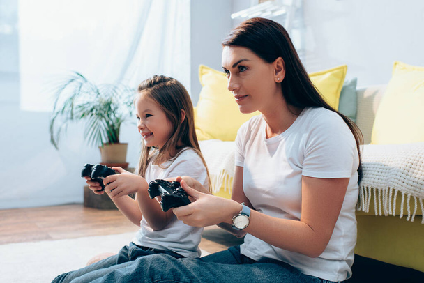 KYIV, UKRAINE - OCTOBER 19, 2020: Smiling mother and daughter looking away while playing with joysticks on floor on blurred background - Photo, Image