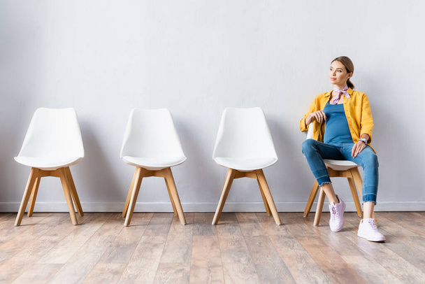 Young pregnant woman looking away while sitting on chair in hall  - Foto, Imagen