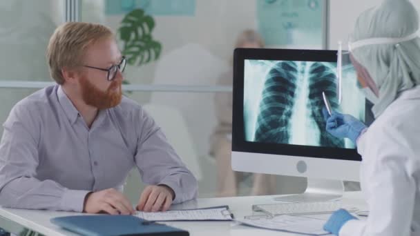 Tıbbi muayenehanede oturan erkek hasta, bilgisayardan göğüs röntgenine bakıyor ve koronavirüs salgını sırasında koruyucu üniformalı müslin kadın doktoru dinliyor. - Video, Çekim
