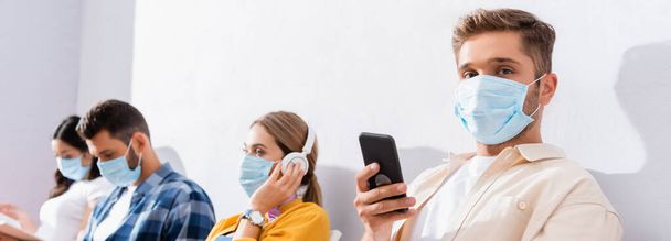 Man in medical mask with smartphone looking at camera near multicultural people on blurred background in queue, banner  - Photo, Image