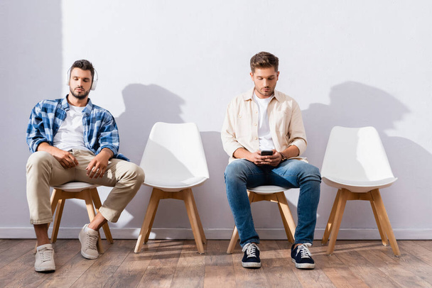 Hombres jóvenes usando auriculares y teléfonos inteligentes en sillas mientras esperan en la sala  - Foto, imagen