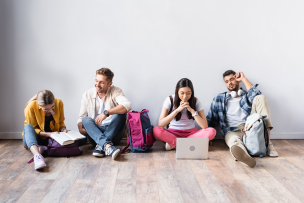 Étudiants multiculturels avec ordinateur portable et livre assis sur le sol dans le hall  - Photo, image