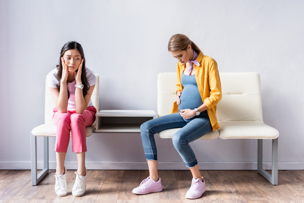 Upset asian woman sitting near pregnant woman while waiting in hall  - Photo, Image