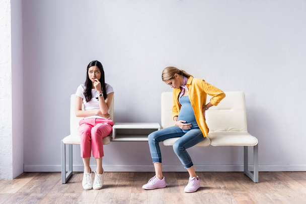 Pregnant woman sitting near worried asian woman in queue  - Photo, Image