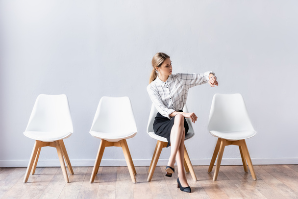 jonge zakenvrouw controleren tijd terwijl zitten met gekruiste benen op stoel  - Foto, afbeelding