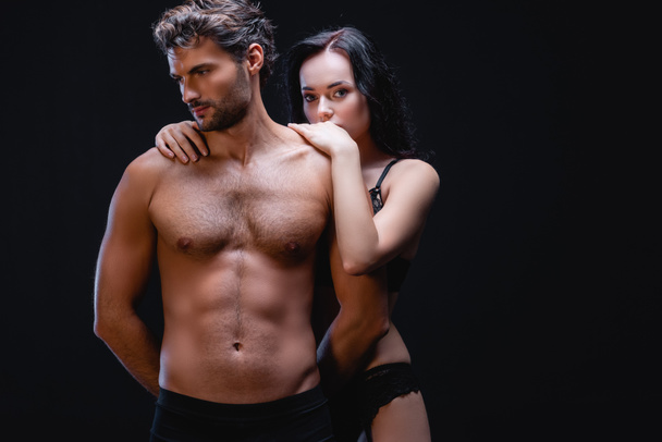 sensual brunette woman embracing shoulders of man with muscular torso while looking at camera isolated on black - Foto, Imagem