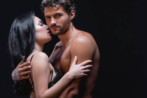 young shirtless man looking at camera while embracing seductive woman in lace bra isolated on black - 写真・画像