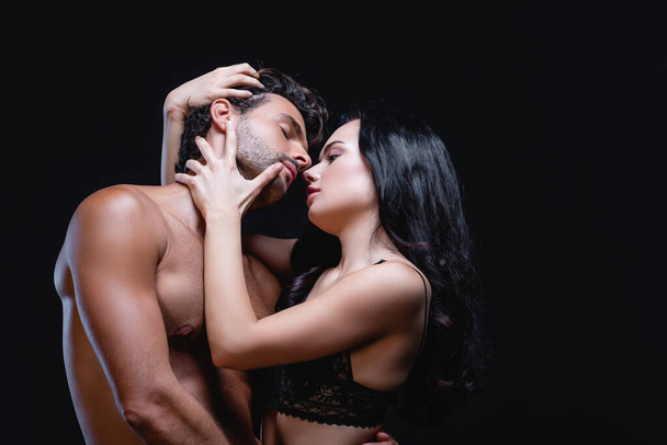 seductive brunette woman embracing head of young shirtless man isolated on black - Foto, immagini