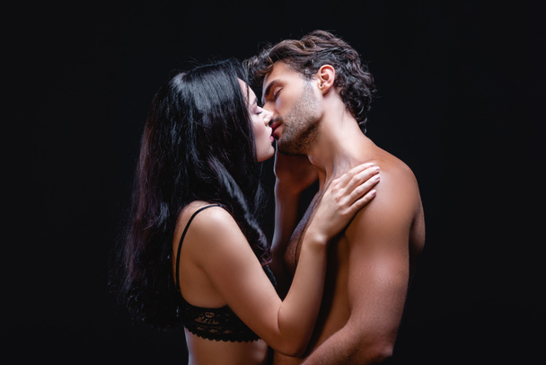 vista lateral da sedutora morena mulher beijando sexy shirtless homem isolado no preto - Foto, Imagem
