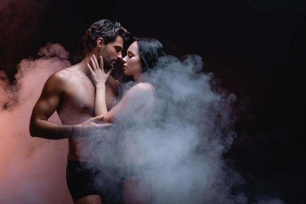 sensual mujer tocando sexy sin camisa hombre mientras que de pie cara a cara sobre fondo oscuro con humo - Foto, Imagen