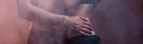 vista recortada del hombre tatuado sin camisa abrazando a la mujer sobre fondo oscuro con humo, pancarta - Foto, imagen