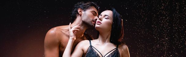 hombre joven sin camisa besar a mujer seductora en sujetador de encaje negro sobre fondo oscuro bajo la lluvia, bandera - Foto, Imagen