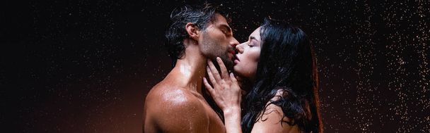 side view of sexy brunette woman and shirtless man kissing with closed eyes on dark background under falling raindrops, banner - Photo, Image