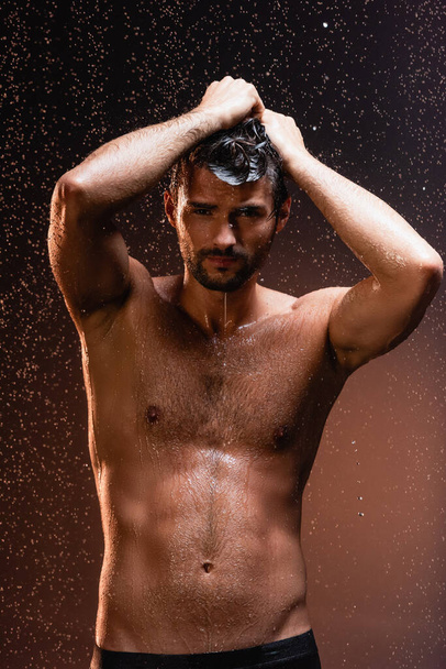 shirtless, muscular man looking at camera under falling raindrops on dark background - Photo, Image
