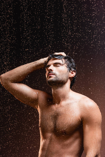 hombre musculoso sin camisa con los ojos cerrados bajo gotas de lluvia cayendo sobre fondo oscuro - Foto, Imagen