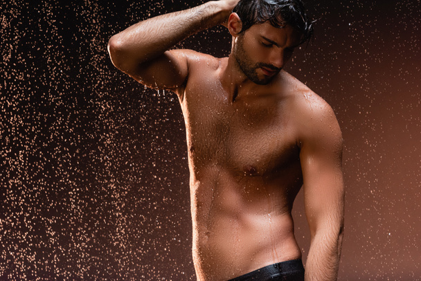 hombre sin camisa con torso muscular posando con la mano en la cabeza bajo la lluvia sobre fondo oscuro - Foto, Imagen