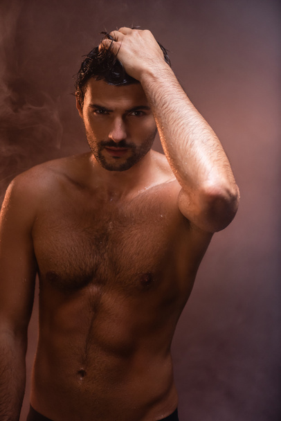 sexy, hombre mojado con torso muscular posando con la mano en la cabeza sobre fondo oscuro con humo - Foto, imagen