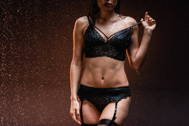 gedeeltelijk uitzicht van sexy vrouw in zwart kant ondergoed en jarretelgordel onder vallende regendruppels op donkere achtergrond - Foto, afbeelding