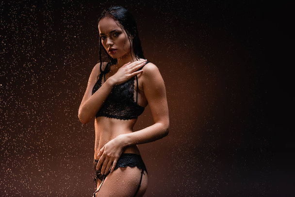 seductive woman in black lace lingerie looking at camera under rain on dark background - Foto, afbeelding