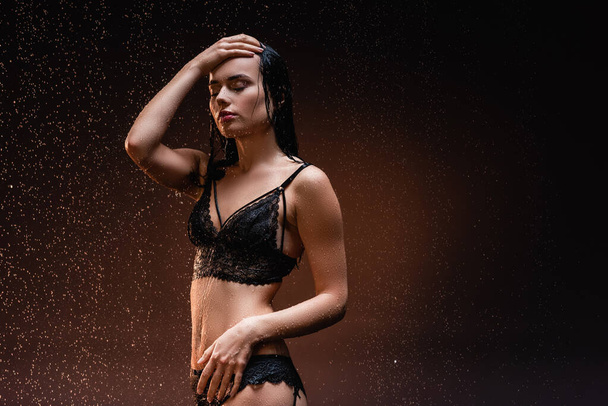 wet, seductive woman with closed eyes posing with hand on head under rain on dark background - Photo, Image