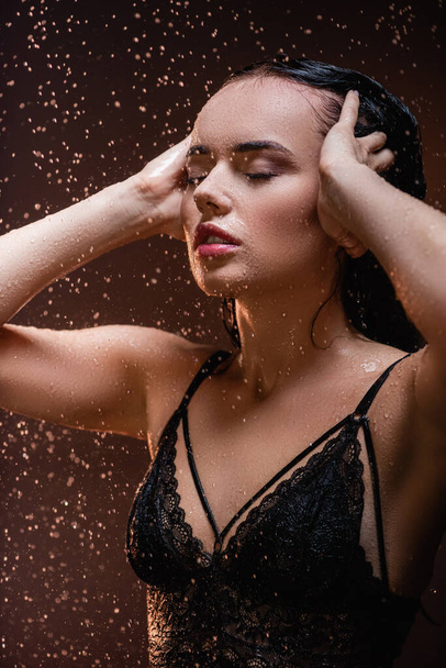 seductive woman in black lace bra posing with closed eyes under falling rain on dark background - Photo, Image