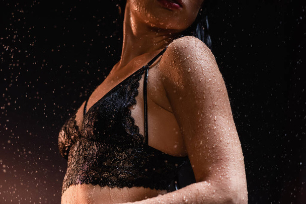 cropped view of wet young woman in black lace bra under rain on dark background - Fotó, kép