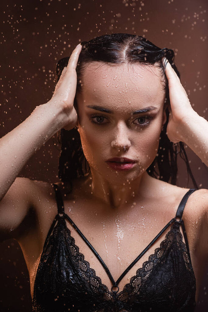 seductive young woman in lace bra looking at camera and touching hair under rain on dark background - Foto, Imagen