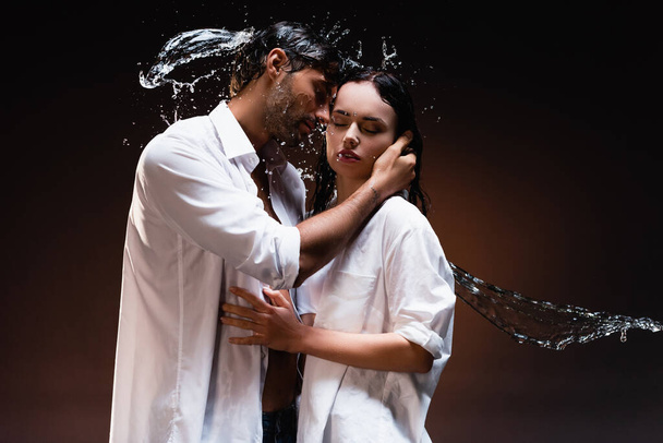 young couple in wet white shirts embracing with closed eyes near water splashes on dark background - Photo, image