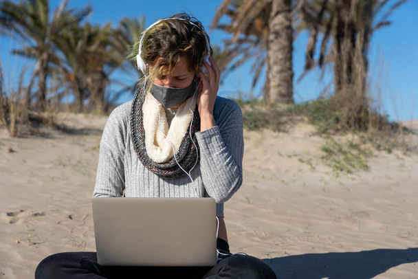 A selective focus shot of female in mask and headphones sitting on a beach with a laptop and working - concept of new normal - Foto, afbeelding
