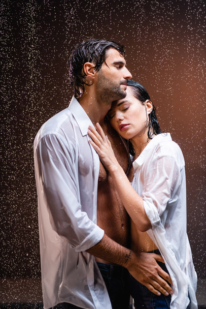 Beyaz ıslak gömlekli genç çift yağmurun altında sarılıyor koyu renk arka planda kapalı gözleri var. - Fotoğraf, Görsel
