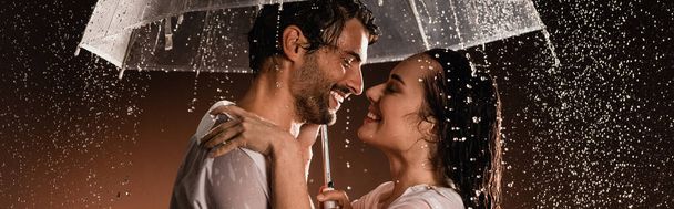 Islak gömlekli mutlu çiftin yan görünüşü yağmurun altında, koyu arkaplanda şeffaf şemsiyeyle yüz yüze duruyor. - Fotoğraf, Görsel