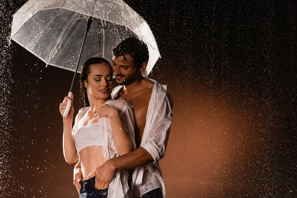 sonriente hombre cogido de la mano en los bolsillos de la mujer sexy de pie bajo la lluvia con paraguas transparente sobre fondo oscuro - Foto, Imagen