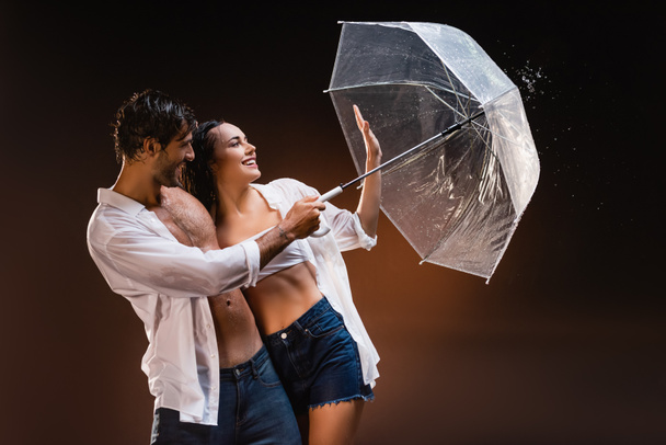 cheerful couple in wet shirts standing with transparent umbrella near water splashes on dark background - Photo, Image