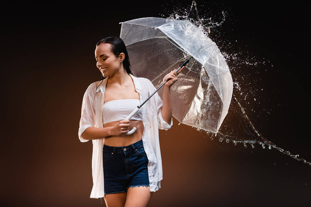 happy woman in white shirt and denim shorts holding umbrella near water splashes - Foto, afbeelding