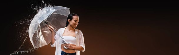 felice, donna bagnata in camicia bianca in piedi con ombrello vicino spruzzi d'acqua su sfondo scuro, banner - Foto, immagini