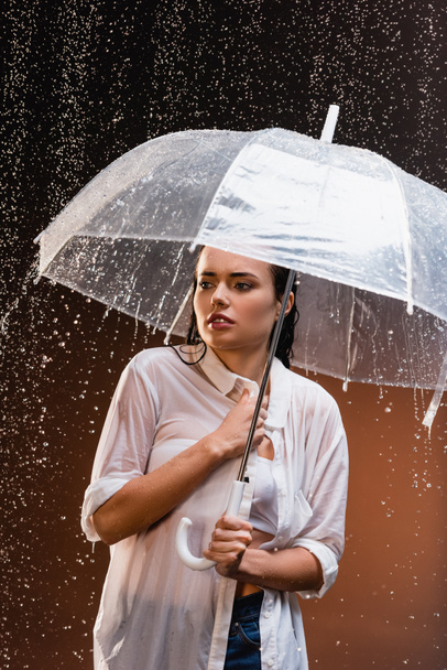 mujer joven en camisa blanca mojada de pie bajo la lluvia con paraguas transparente sobre fondo oscuro - Foto, Imagen
