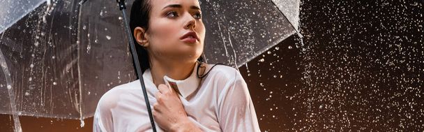 mujer mojada mirando hacia otro lado mientras está de pie con lluvia bajo paraguas transparente sobre fondo oscuro, pancarta - Foto, Imagen
