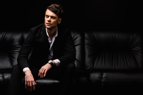 confident man in formal wear sitting on sofa isolated on black background  - Fotografie, Obrázek