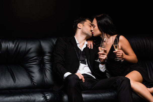 casal sexy beijando e segurando óculos com champanhe enquanto sentado no sofá isolado no preto  - Foto, Imagem