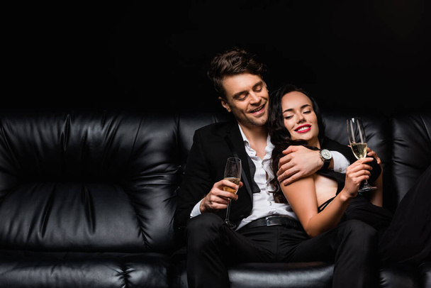 smiling couple holding glasses of champagne while resting on sofa isolated on black - Foto, Imagem