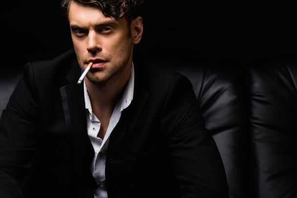 confident man in formal wear smoking cigarette and looking at camera isolated on black  - Фото, изображение