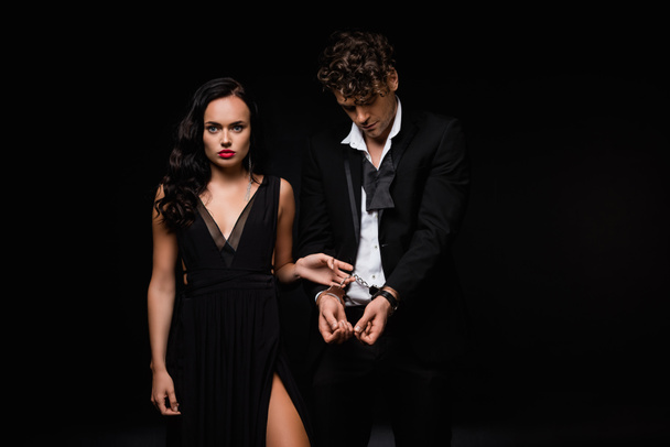 sexy woman in dress holding handcuffs on submissive man isolated on black - Fotó, kép