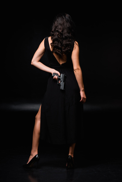 back view of dangerous woman holding gun behind back on black  - Φωτογραφία, εικόνα