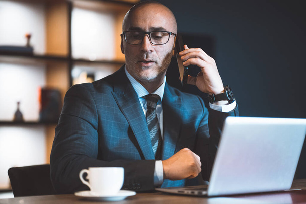 Answering a phone call, a businessman took a break from work and looks at a cup of coffee near a laptop.. Handsome bold man working on laptop - Foto, imagen