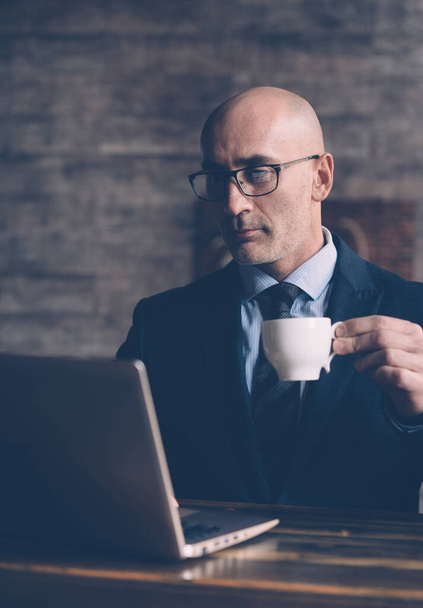 Taking a coffee break and looking through business correspondence, a businessman holds a cup of coffee in his hand and looks into the laptop monitor wearing eye glasses. Business concept. Toned image - Photo, image