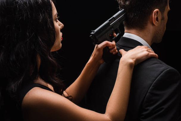dangerous woman holding gun behind man in suit isolated on black  - 写真・画像