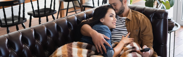 joven hombre sosteniendo mando a distancia y abrazando novia sonriente viendo la televisión bajo una manta acogedora, pancarta - Foto, imagen
