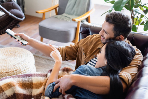 cheerful man holding remote controller near brunette woman pointing with finger while watching movie at home - Photo, Image
