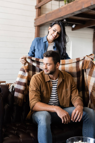 happy woman covering boyfriend with warm plaid blanket at home - Foto, afbeelding