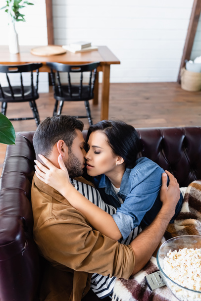 young couple hugging and kissing on leather sofa near wooden table and chairs on blurred foreground - Fotografie, Obrázek